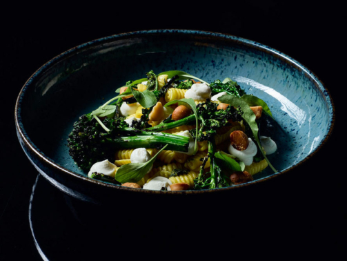 Fundo escuro com um prato azul centralizado com gnocchetti sardi servido com pesto de rúcula, creme de macadâmia e brócolis tostado. Faz parte do novo menu do Libreria Augusto.