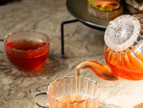 Imagem mostra um bule de vidro com chá sendo colocado em uma xícara.