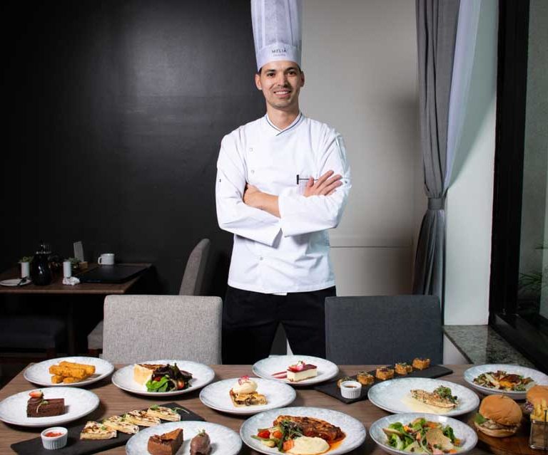 Imagem mostra o Chef Rodrigo Leite em frente a uma mesa com diversos pratos de comida